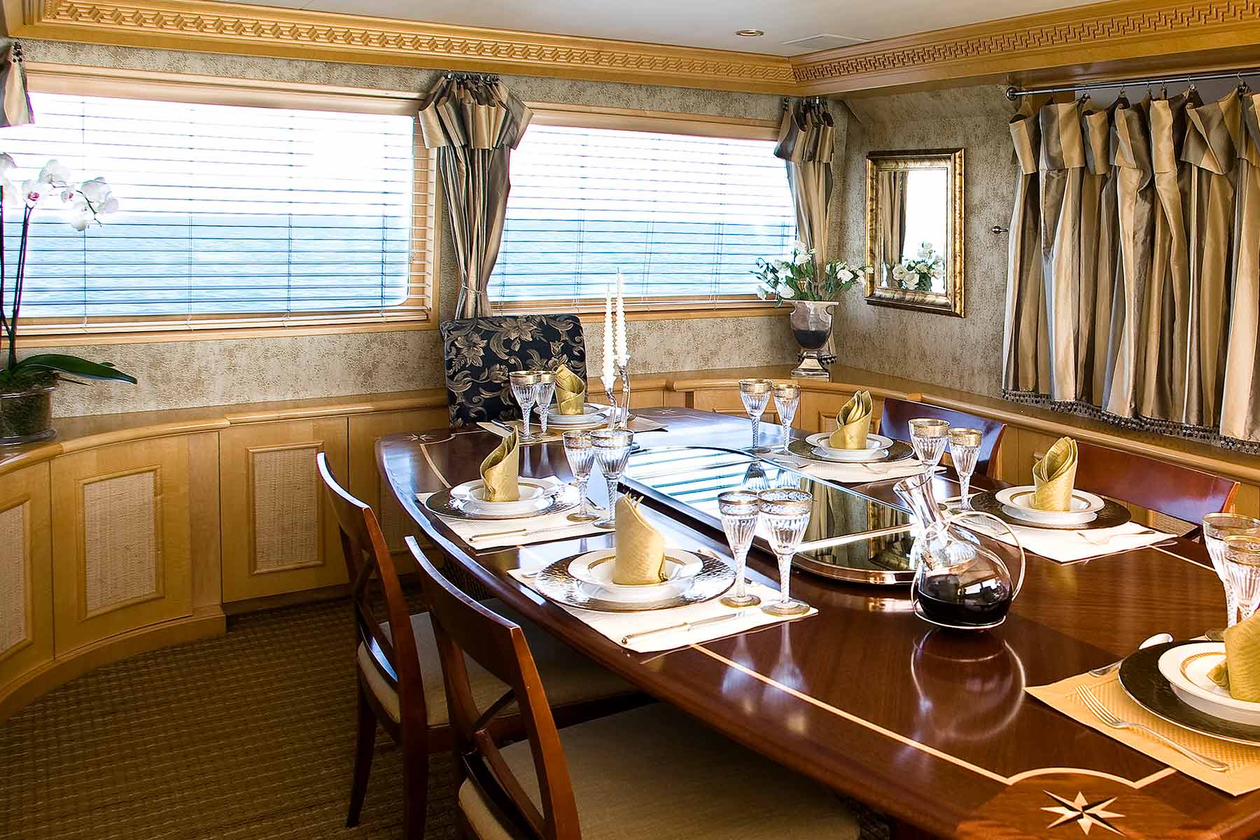 Elegant yacht dining area with a polished wooden table set with gold-rimmed glasses, folded gold napkins, a wine decanter, and floral decorations.
