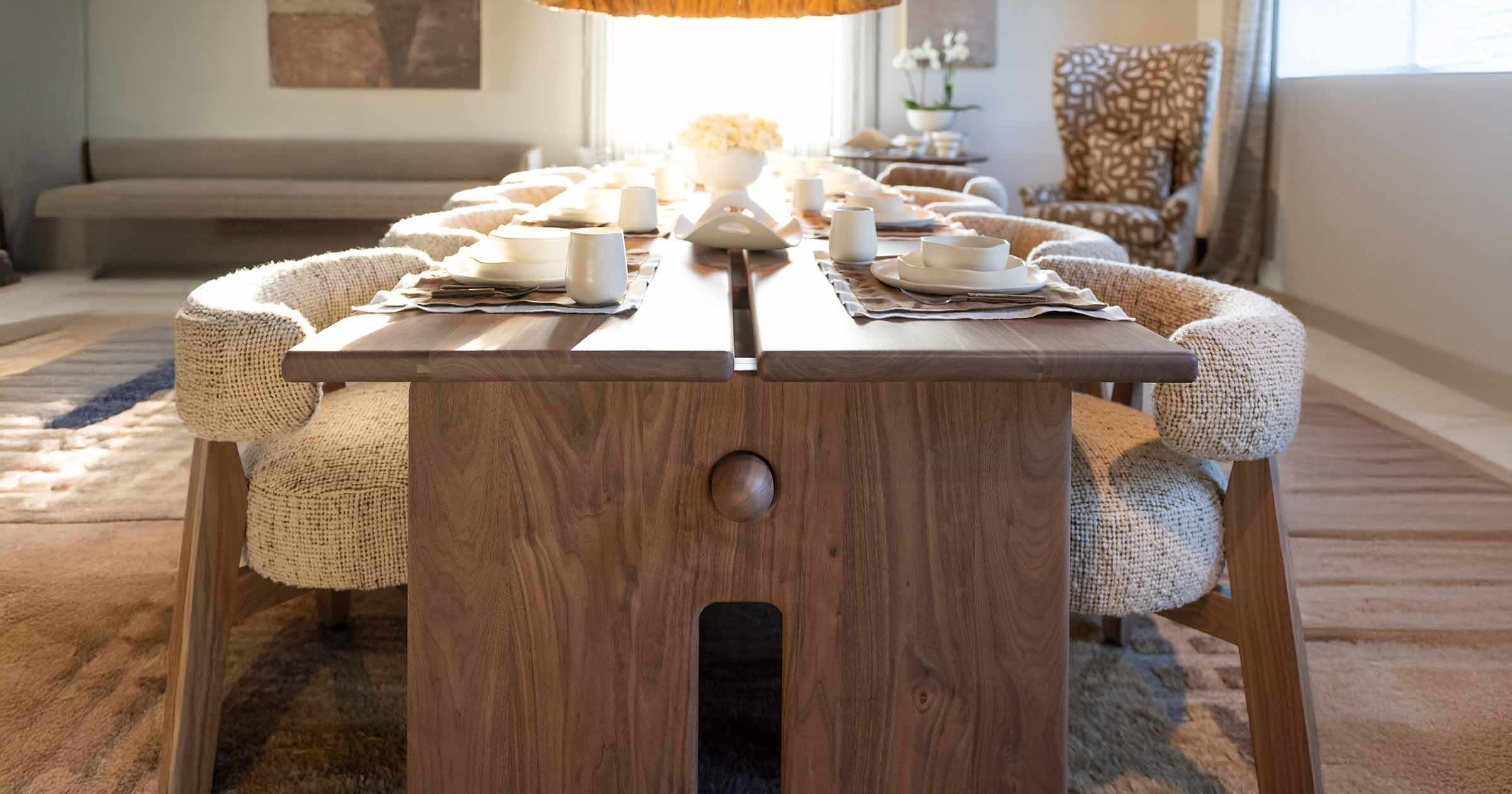 Wooden dining table with split plank design set with white tableware and surrounded by textured beige chairs in a sunlit modern dining room.
