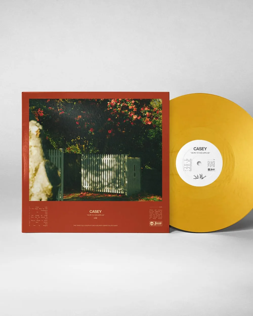 Yellow vinyl record partially inserted in a red album cover featuring a white picket fence and blooming flowers, titled 'Casey - How to Disappear'.