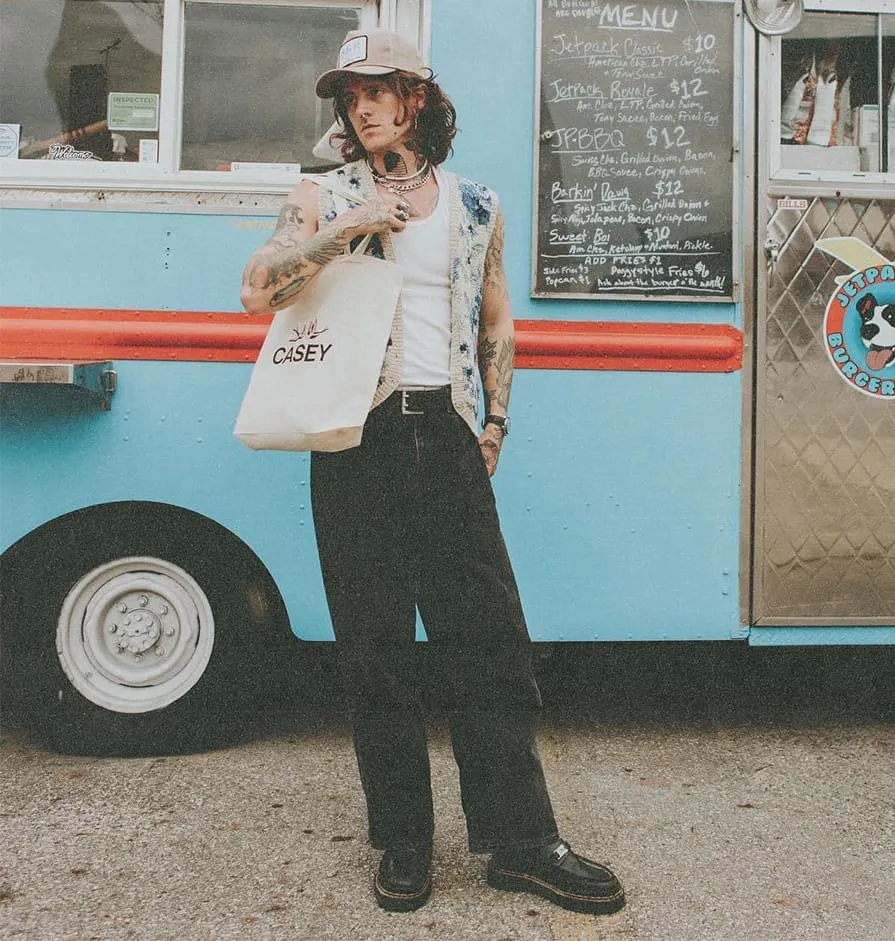 Tattooed person in a cap and vest holding a white tote bag that says CASEY, standing in front of a blue food truck with a menu board.