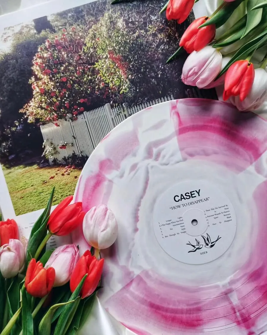 Pink and white marbled vinyl record labeled 'CASEY HOW TO DISAPPEAR' surrounded by red and pink tulips on a surface with a photo of a garden and white picket fence.