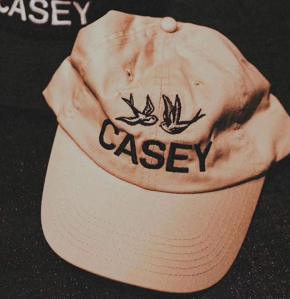 Beige baseball cap with two embroidered birds and the name CASEY in black letters.