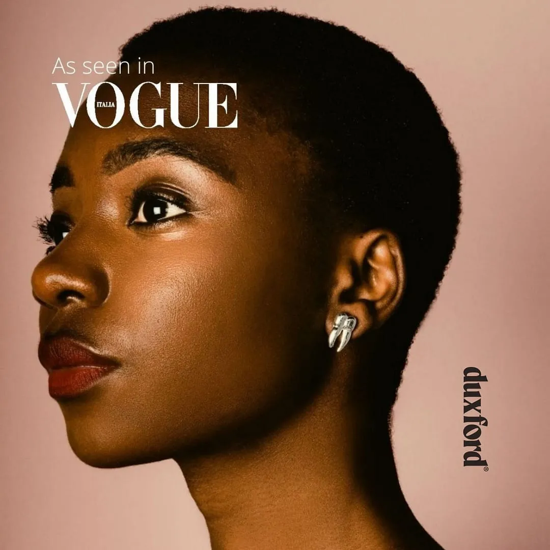 Close-up profile of a woman with short hair wearing silver geometric earrings against a pink background with 'As seen in Vogue Italia' text.
