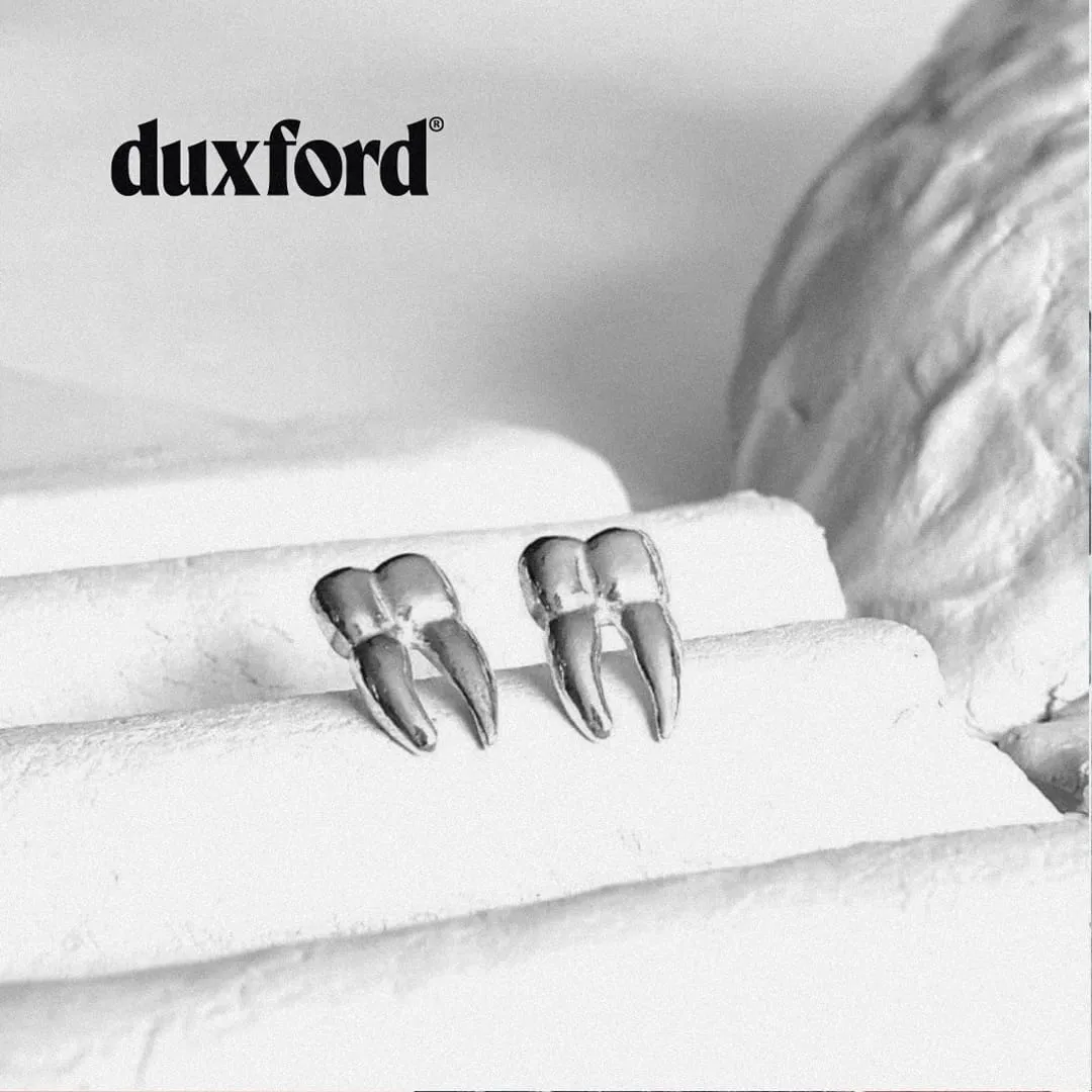 Two shiny metal tooth-shaped earrings placed on white textured blocks with a blurred background.