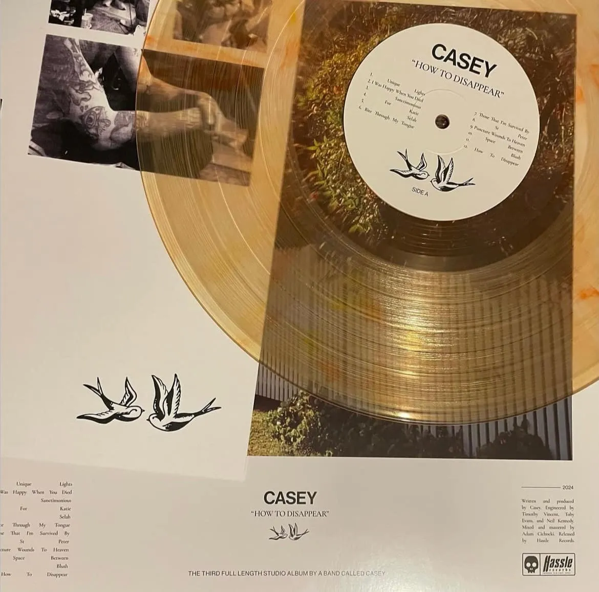 Clear vinyl record titled 'HOW TO DISAPPEAR' by Casey, resting on an album cover with two illustrated birds and tattooed arms visible.