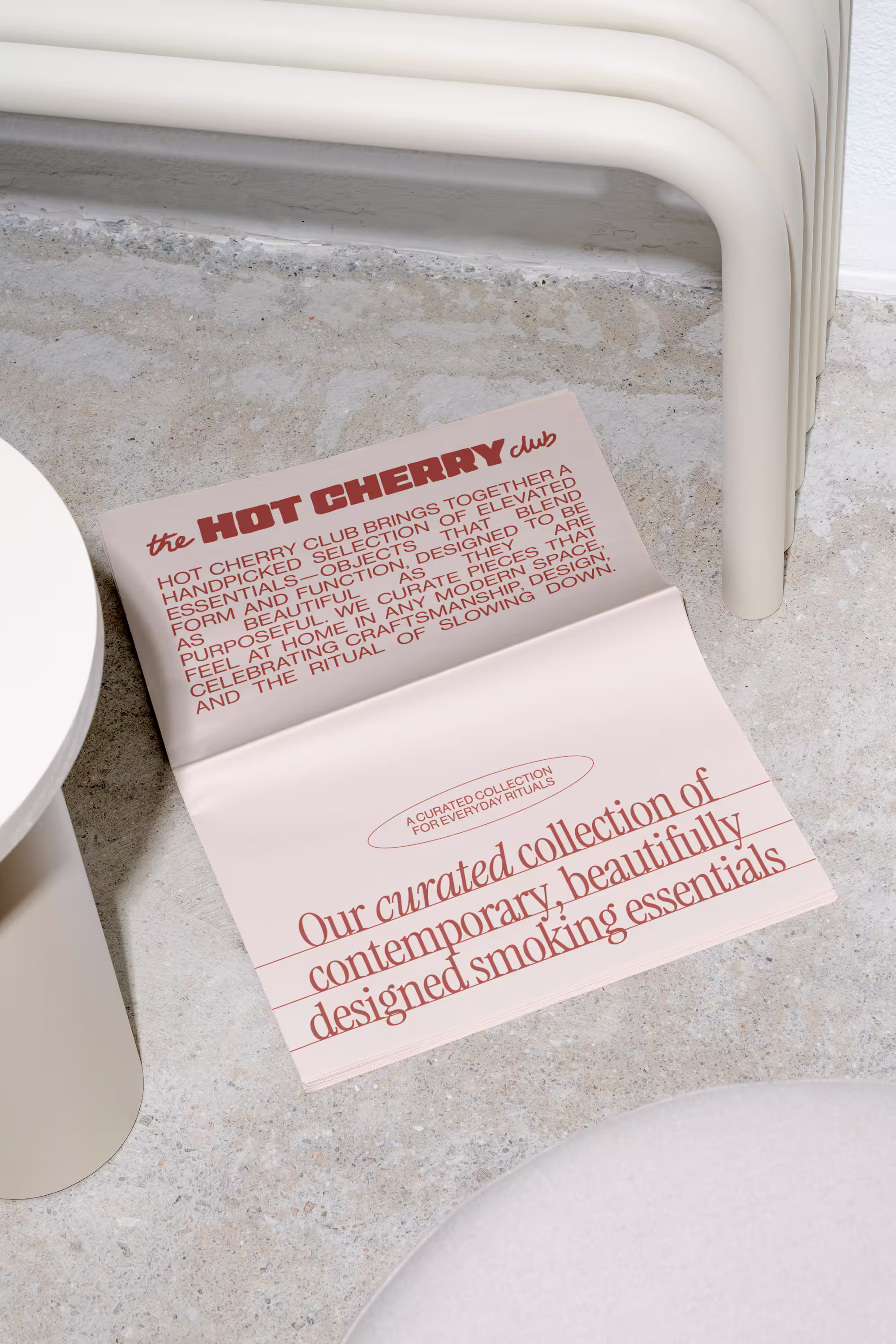 An open booklet on a concrete floor with text about The Hot Cherry Club's curated collection of contemporary, beautifully designed smoking essentials.