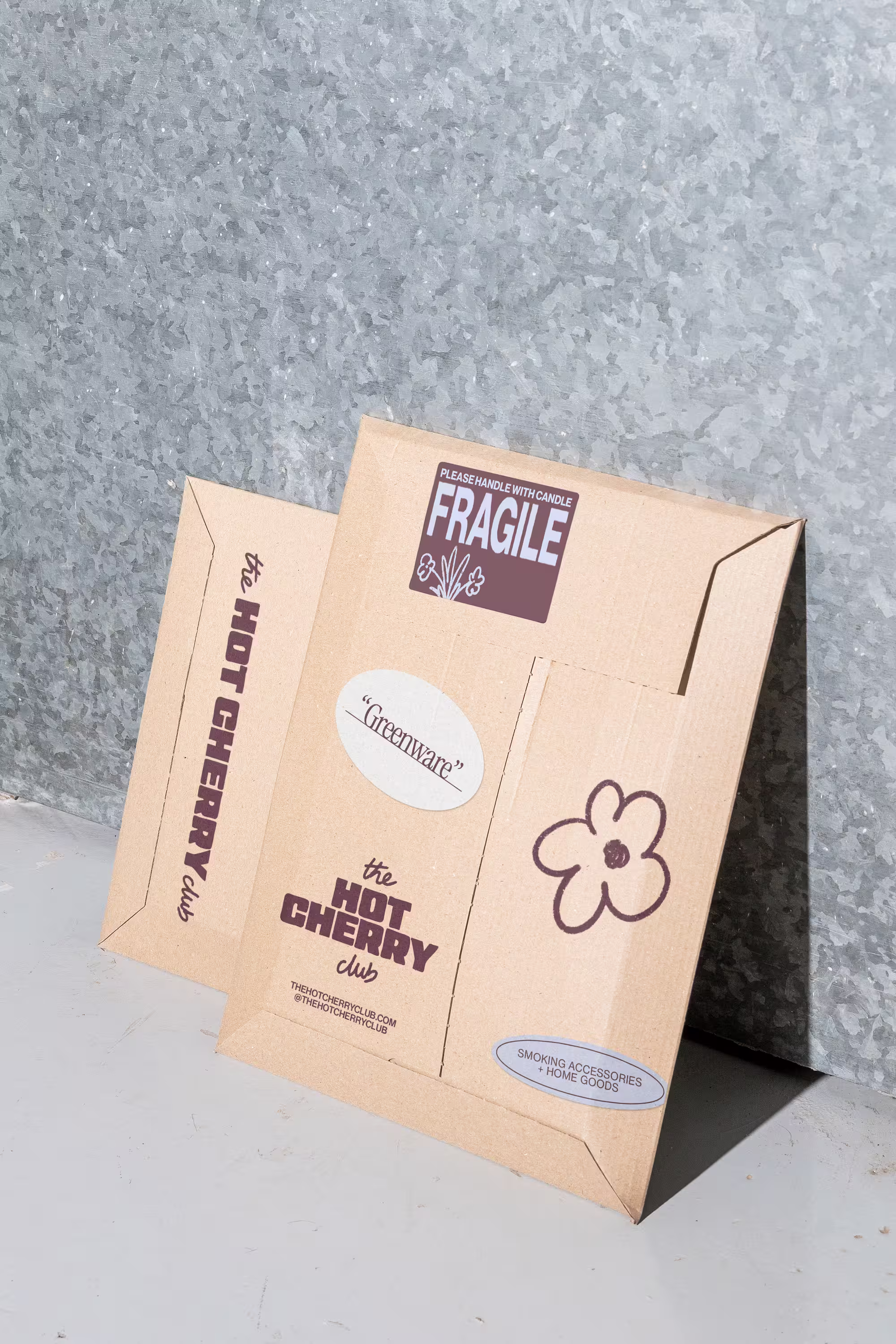Two cardboard boxes leaning against a textured metal wall, labeled with 'the HOT CHERRY club', 'FRAGILE please handle with care', 'Greenware', and 'Smoking Accessories + Home Goods'.