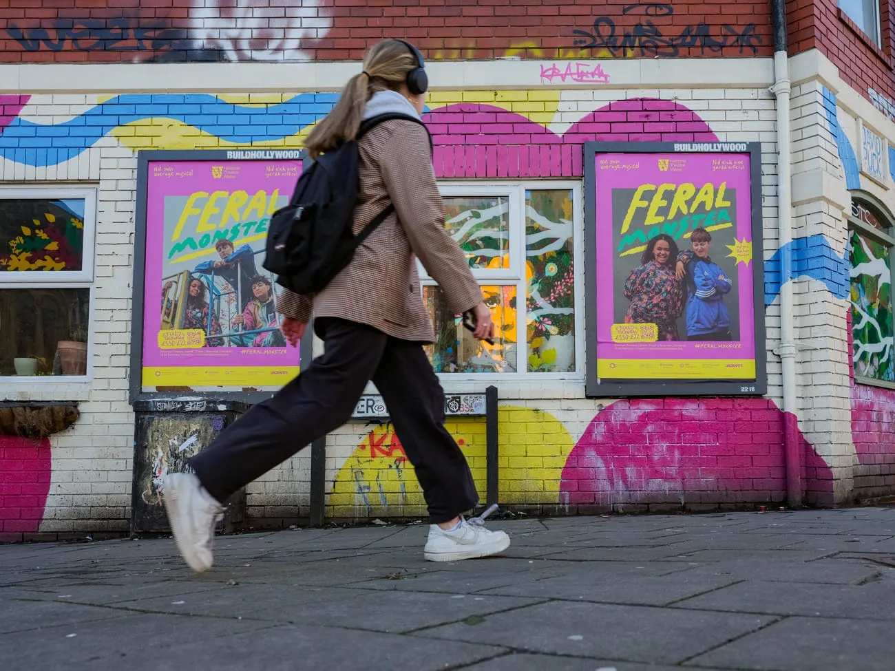 Person wearing headphones, a brown jacket, black pants, and white sneakers walking past a colorful brick wall with two posters advertising 'FERAL MONSTER'.