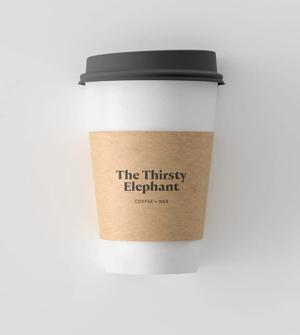 White disposable coffee cup with a black lid and a brown sleeve labeled 'The Thirsty Elephant Coffee + Bar'.