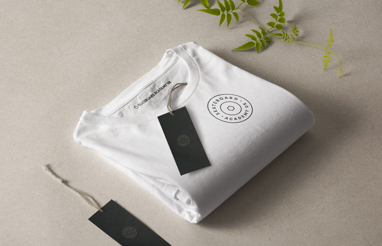 Folded white t-shirt with a black circular Skateboard Academy UK logo and attached black tags on a beige surface with green leaves nearby.