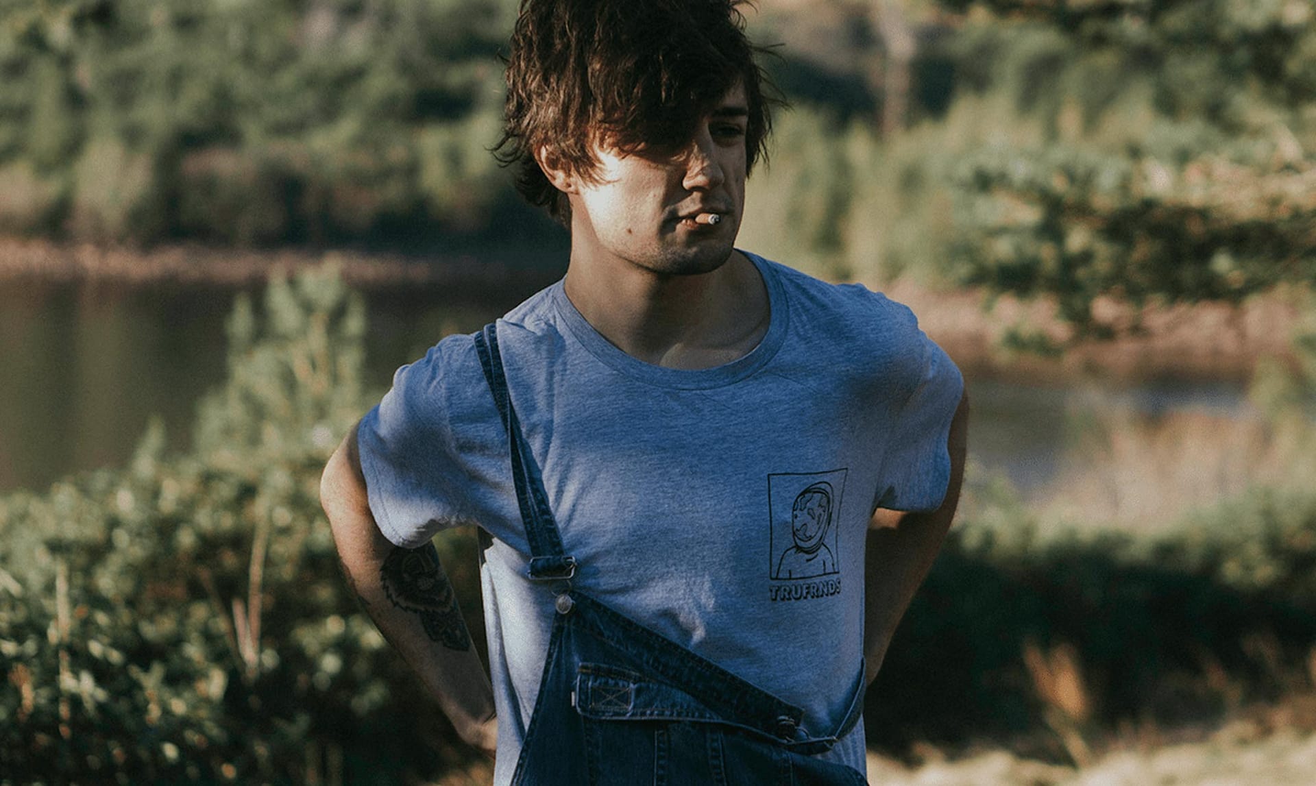 Young man with tousled hair smoking a cigarette, wearing a gray T-shirt with a cartoon face and the word TRUFRNDS, and denim overalls outdoors near greenery and water.