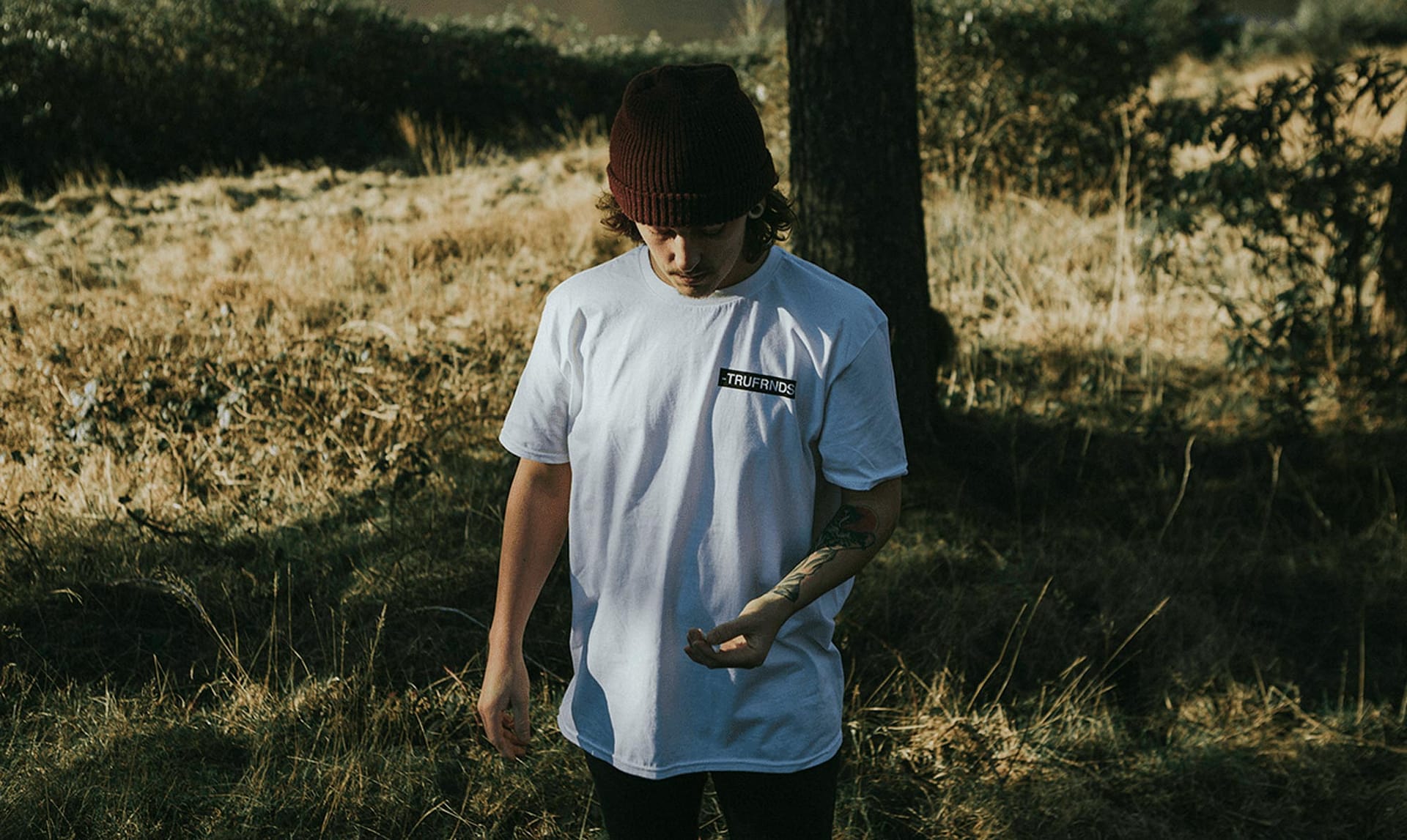 Young man wearing a white TRUFRNDS t-shirt and a maroon beanie standing outdoors in a grassy area with trees.