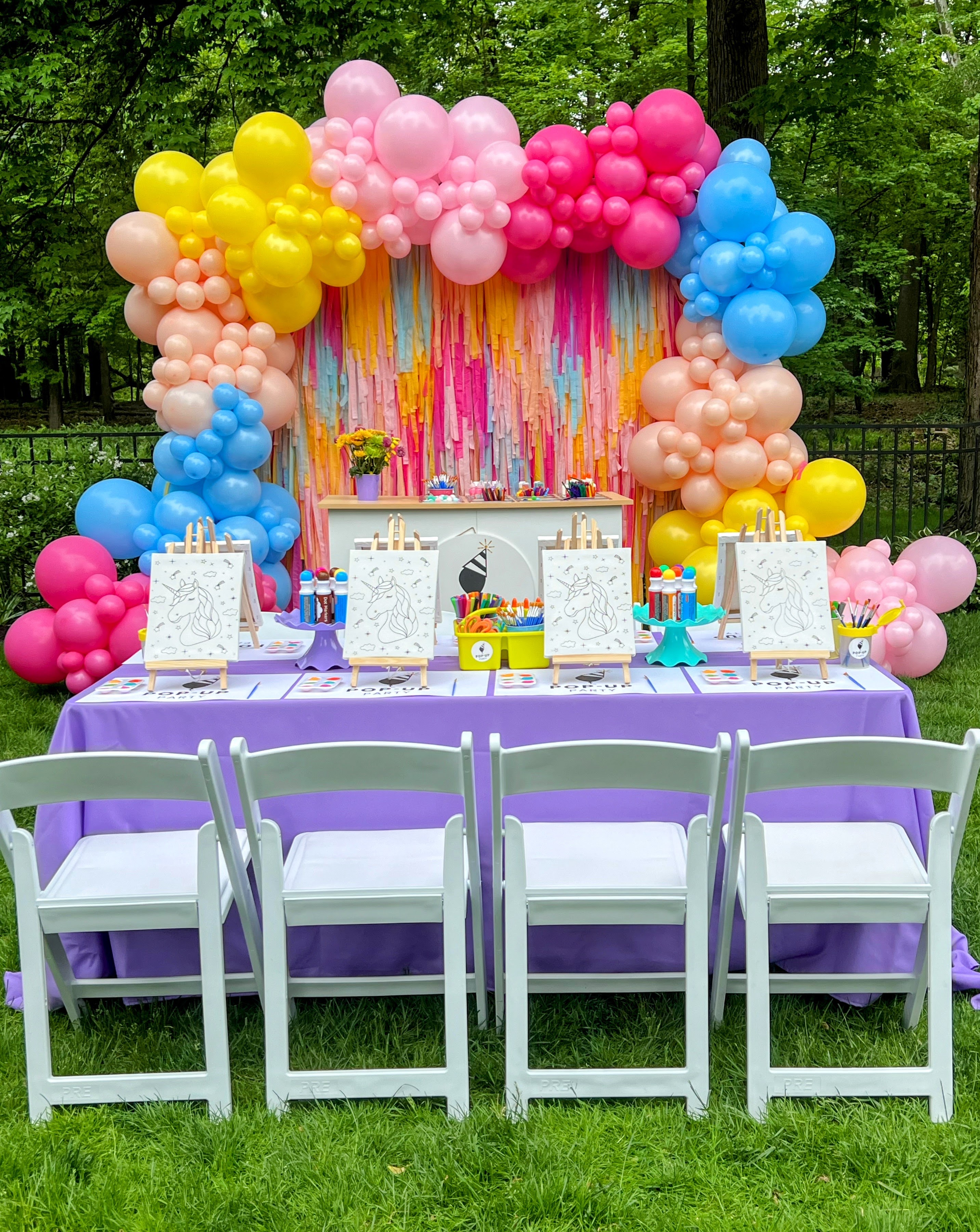 Outdoor kids' painting party setup with unicorn canvases, art supplies, and colorful balloon arch.
