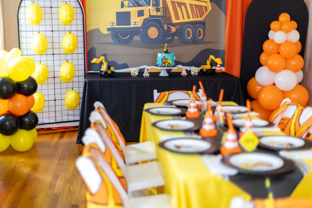 Construction-themed party setup with toy trucks, traffic cones, hard hat balloons, and a blue cake on black tablecloth.