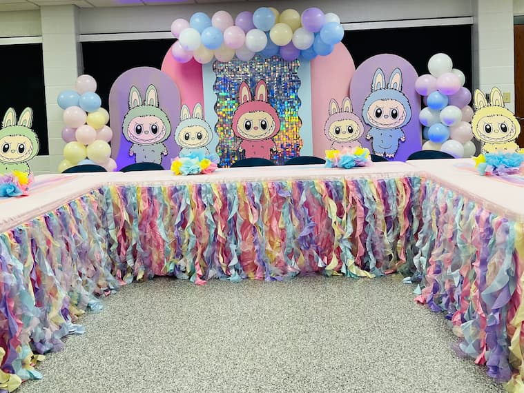 Colorful party table with pastel streamers, balloons, and cute bunny character cutouts on a decorated backdrop.