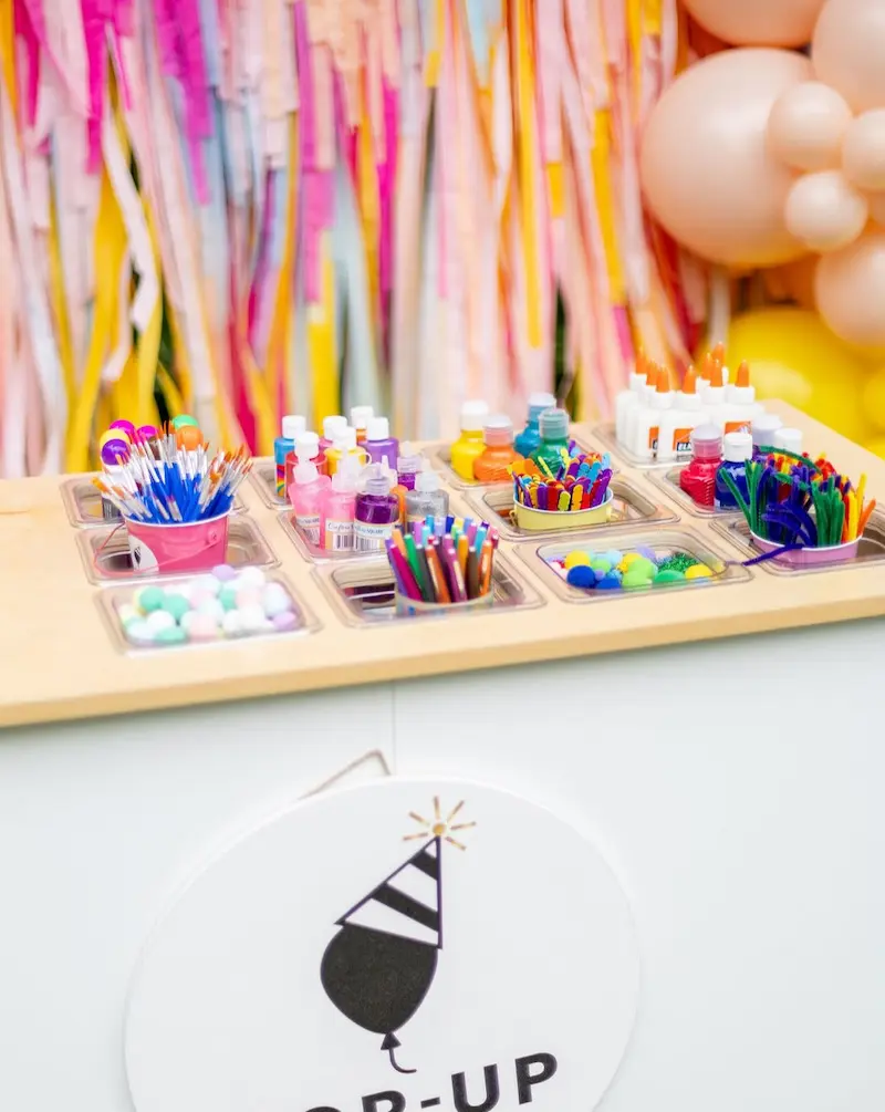 Table with organized art supplies including paintbrushes, paint bottles, glue, markers, pom-poms, and pipe cleaners.