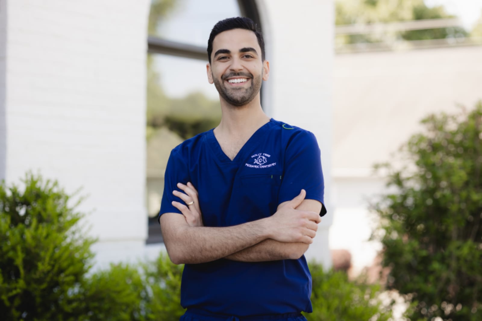 Male dentist in blue scrubs smiling with arms crossed in office setting