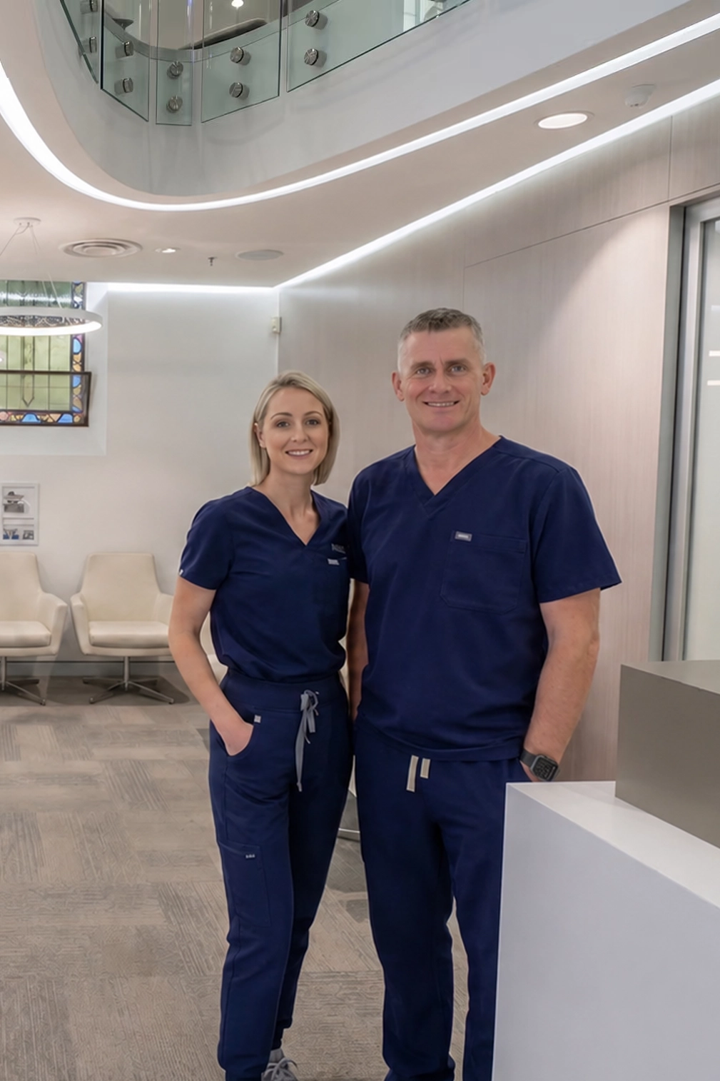 Dr Nathan Connelly and Nicole Starbuck-Connelly standing in their clinic reception area wearing navy scrubs, with white lounge chairs and soft lighting in the background.