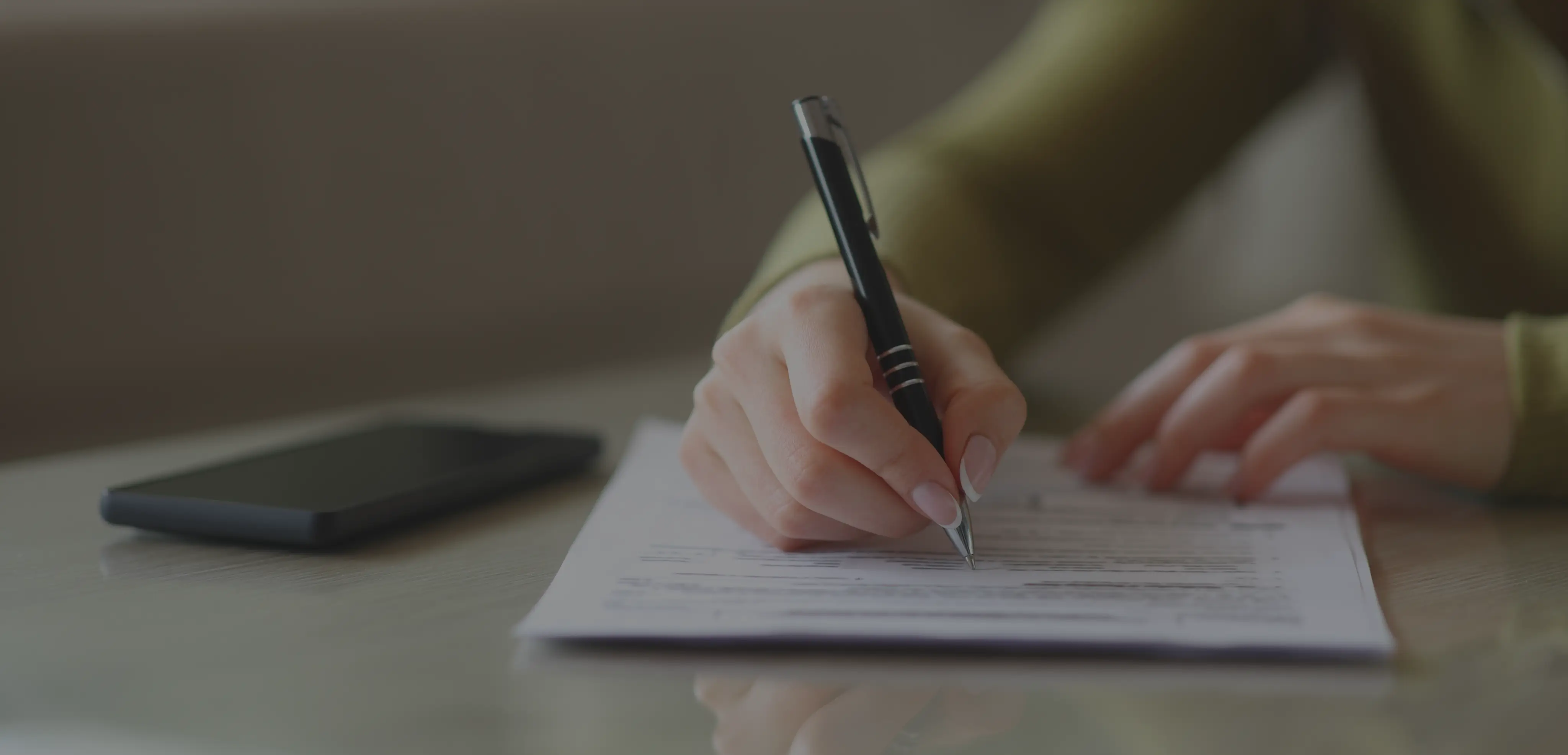 Person filling out a form with a pen on a table next to a black smartphone.