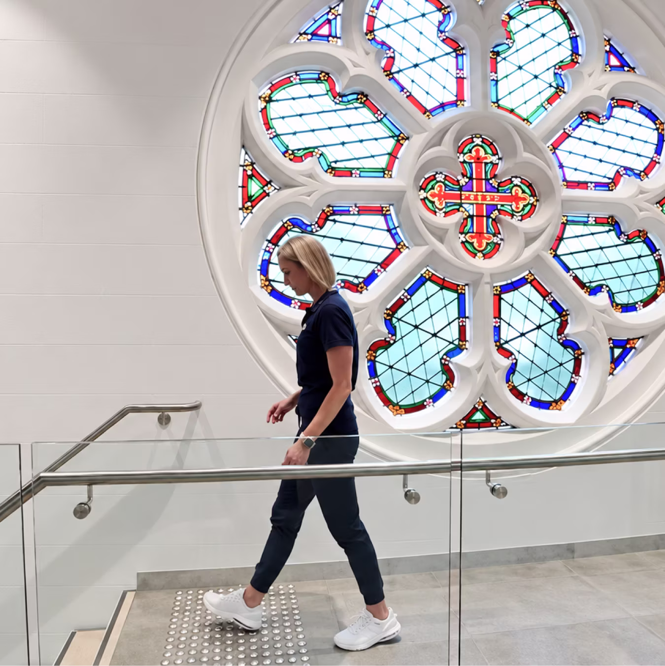 A woman in navy blue clothes walking down indoor stairs with a glass railing, beneath a large circular stained glass window with red, blue, green, and white patterns.