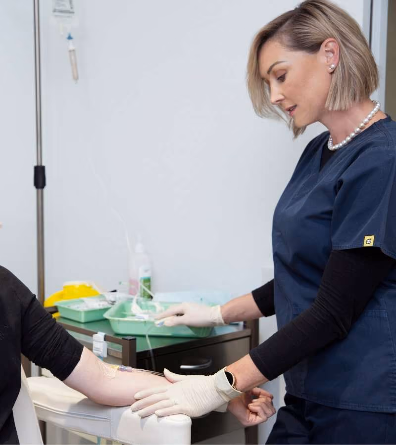 A nurse in navy scrubs and gloves monitors an IV infusion on a patient's arm during a medical procedure in a clinic.