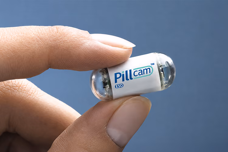 A PillCam SB 2 capsule endoscopy device, shaped like a large vitamin, resting on a polished timber surface.