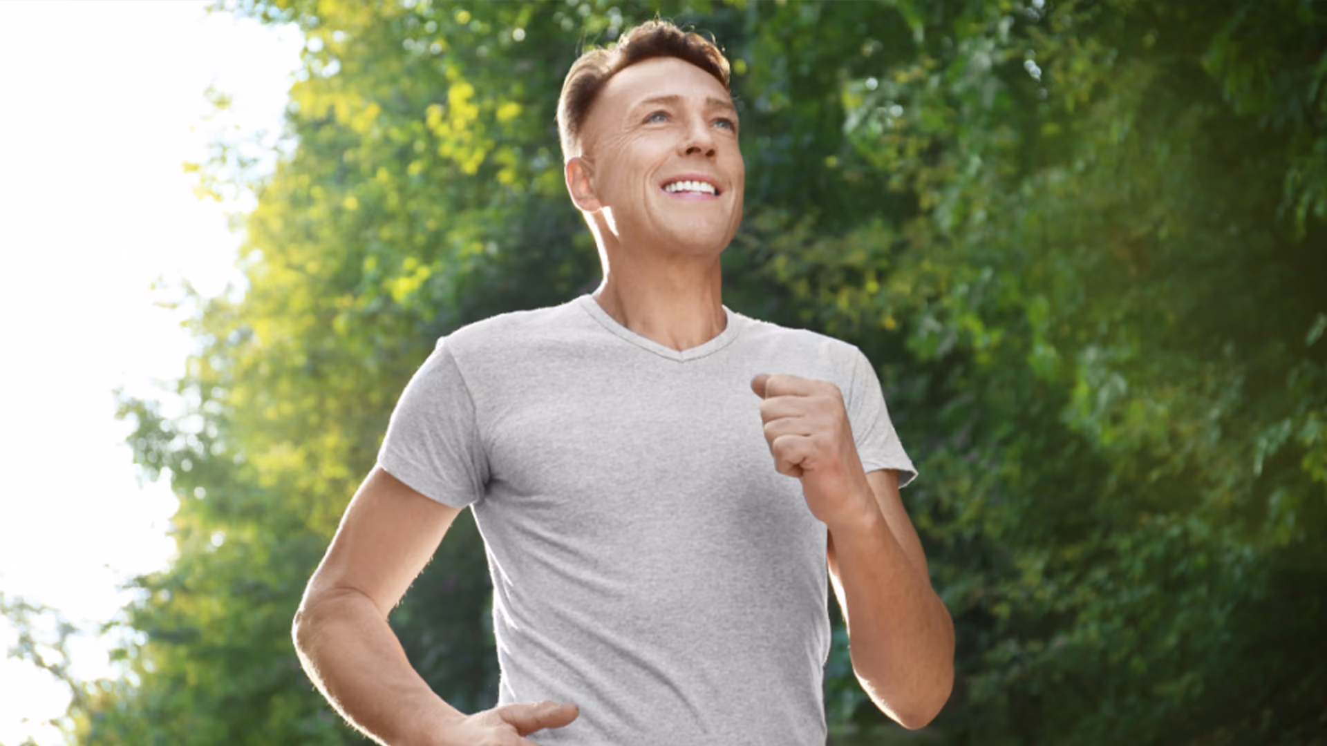 A smiling man in a grey t-shirt jogs outdoors, looking up with a cheerful expression against a background of green trees.