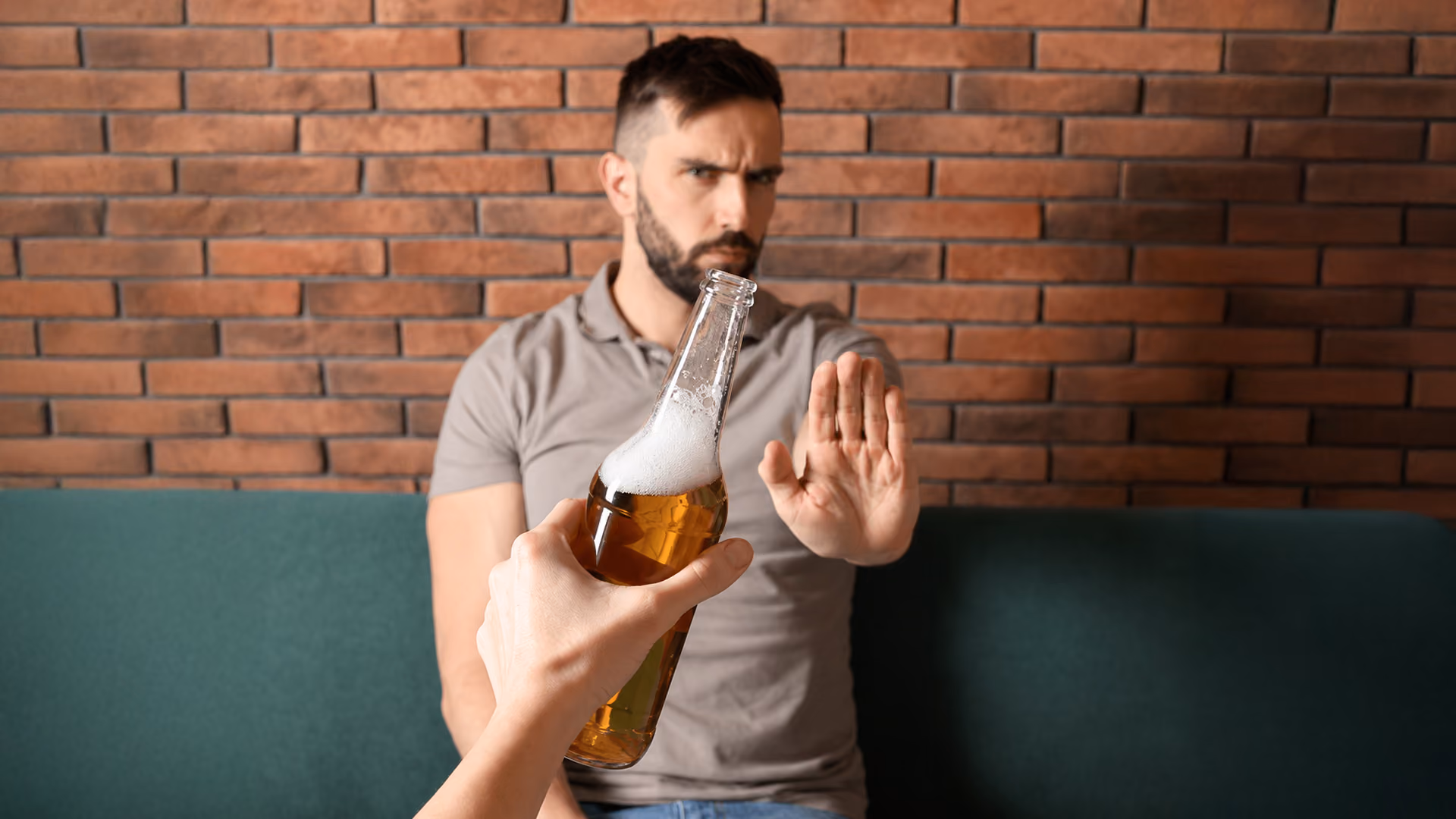 A man makes a stop gesture with his hand, refusing a bottle of beer offered to him, set against a brick wall background.