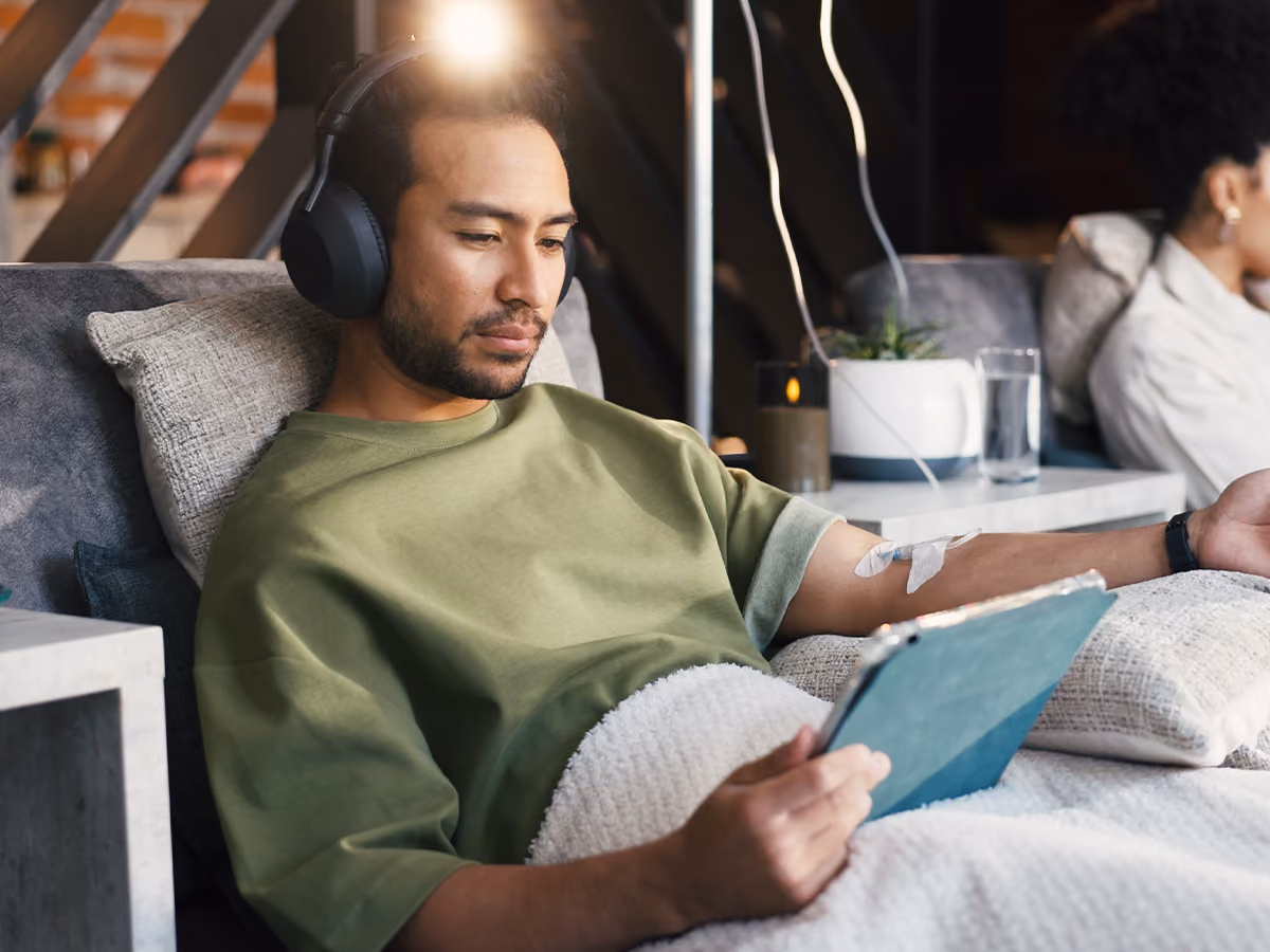 Person relaxing in a comfortable chair while receiving a vitamin infusion through an IV cannula, using a tablet during treatment.