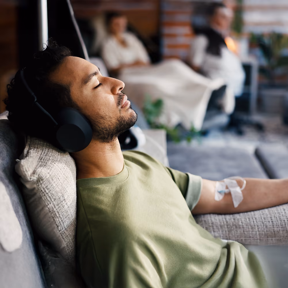 Person relaxing in a comfortable chair with an IV cannula in their arm during a vitamin infusion, wearing headphones while receiving treatment.