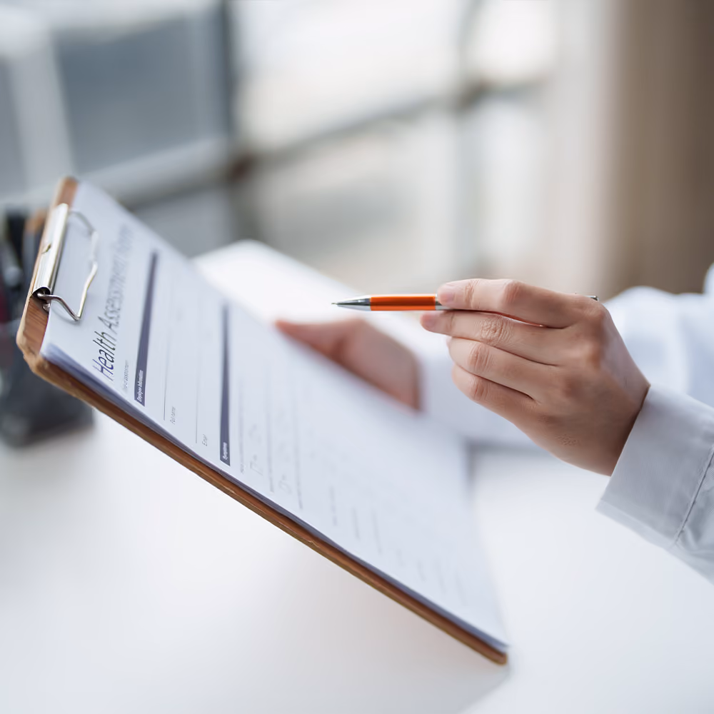 Healthcare professional reviewing a patient health assessment form on a clipboard while discussing additional assessment requirements.