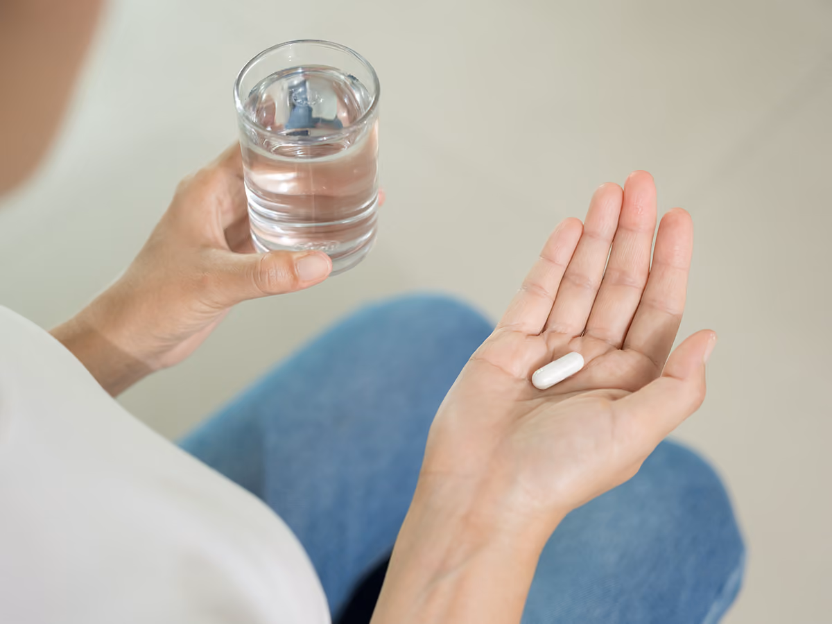 A person holds a white medication capsule in one hand and a glass of water in the other, preparing to take the medication.