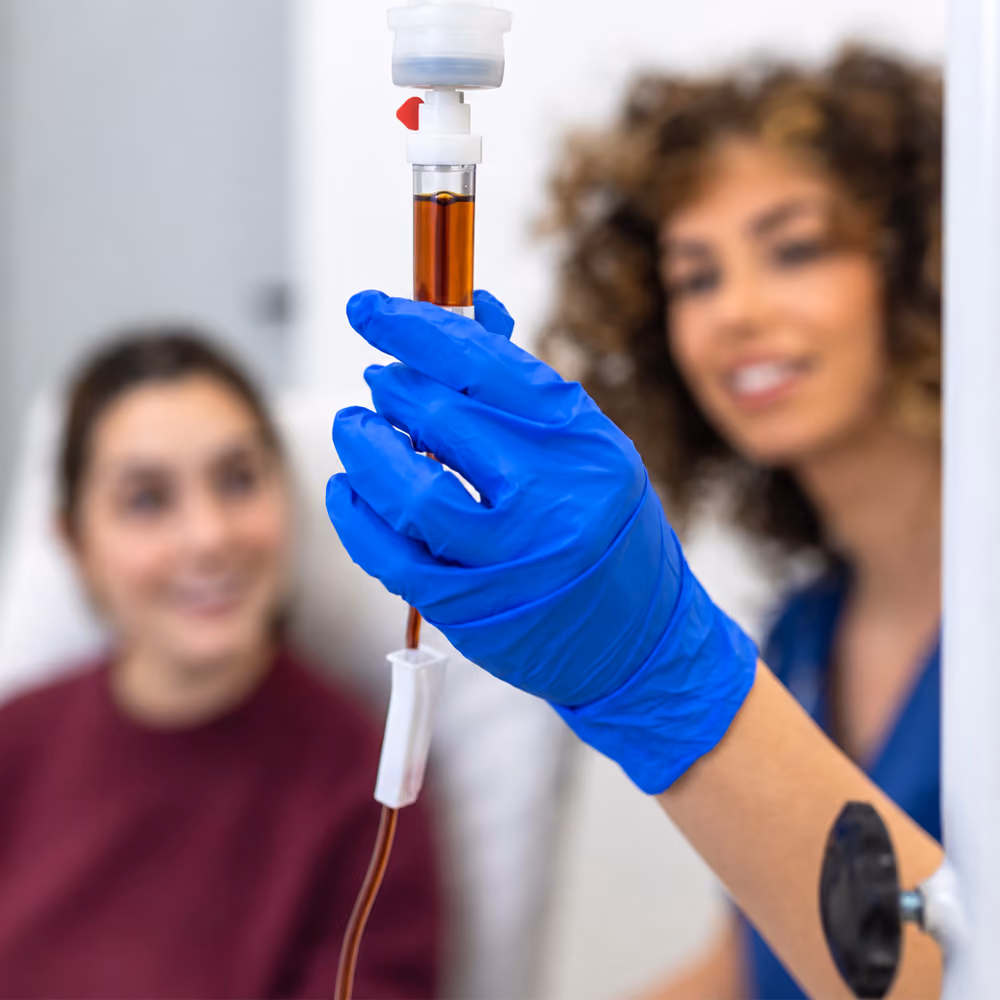 Healthcare professional adjusting an IV drip containing iron infusion fluid while a patient sits in the background.