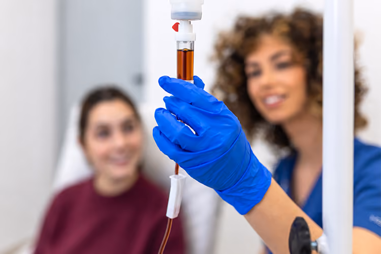 Healthcare professional adjusting an IV drip containing iron infusion fluid while a patient sits in the background.