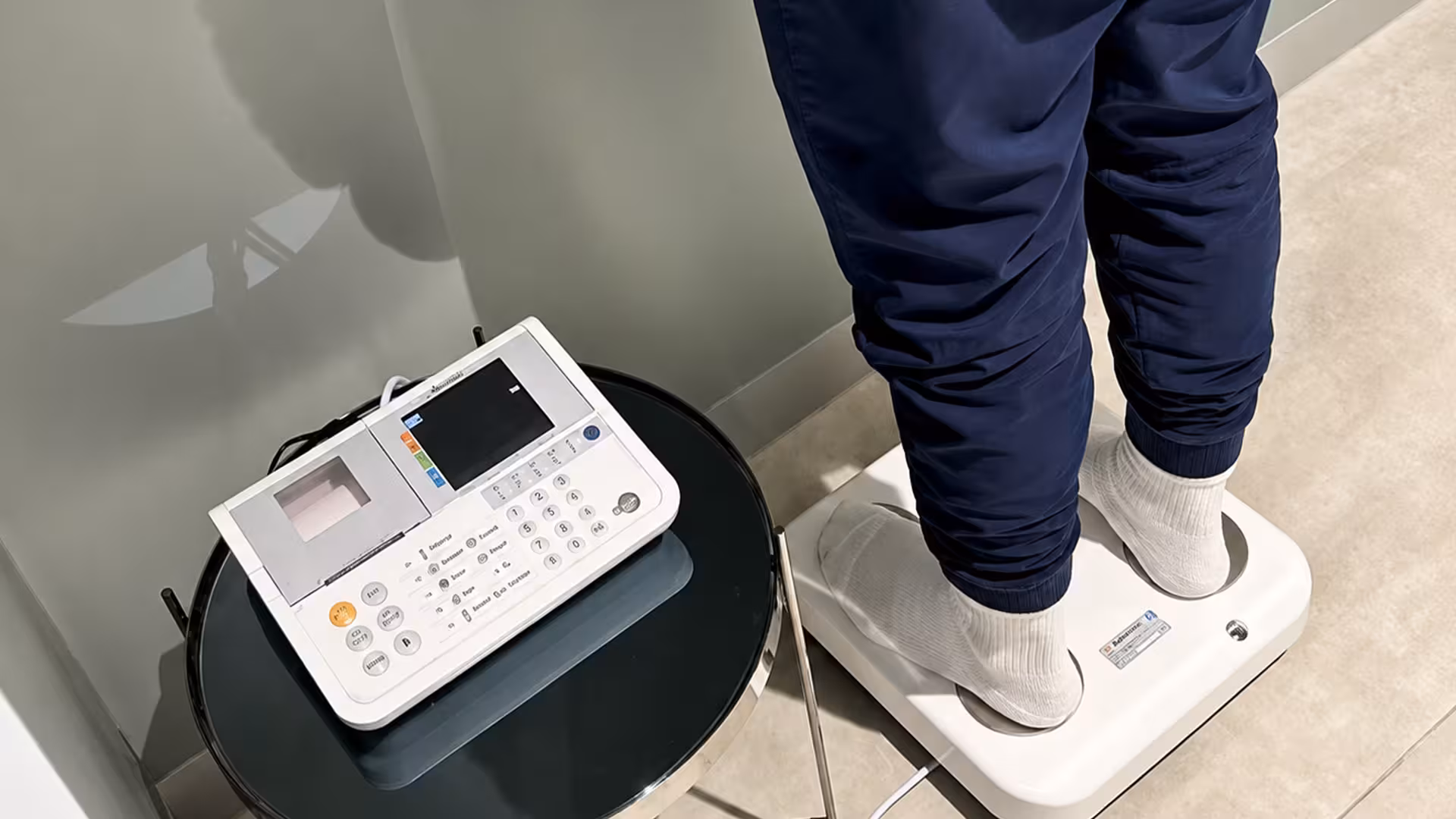 A person in socks stands on a body composition scale connected to a control console with a keypad on a small side table.