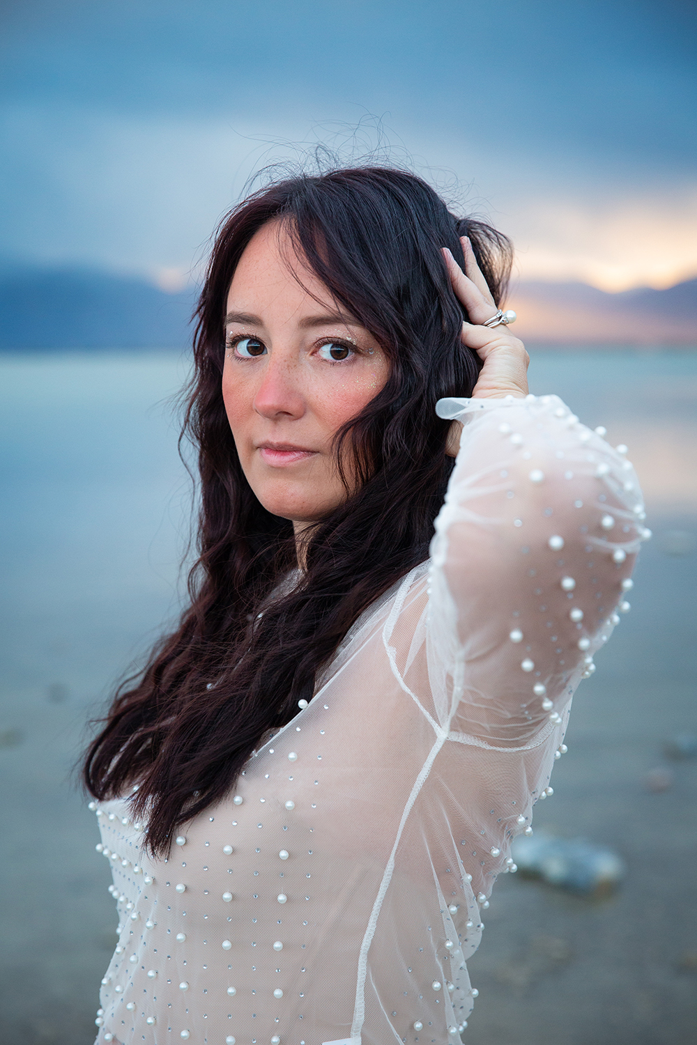 Elaine- girl long dark hair looking into camera. Background mountains at sunrise with blue, purple and pink sky and bright blue water. 