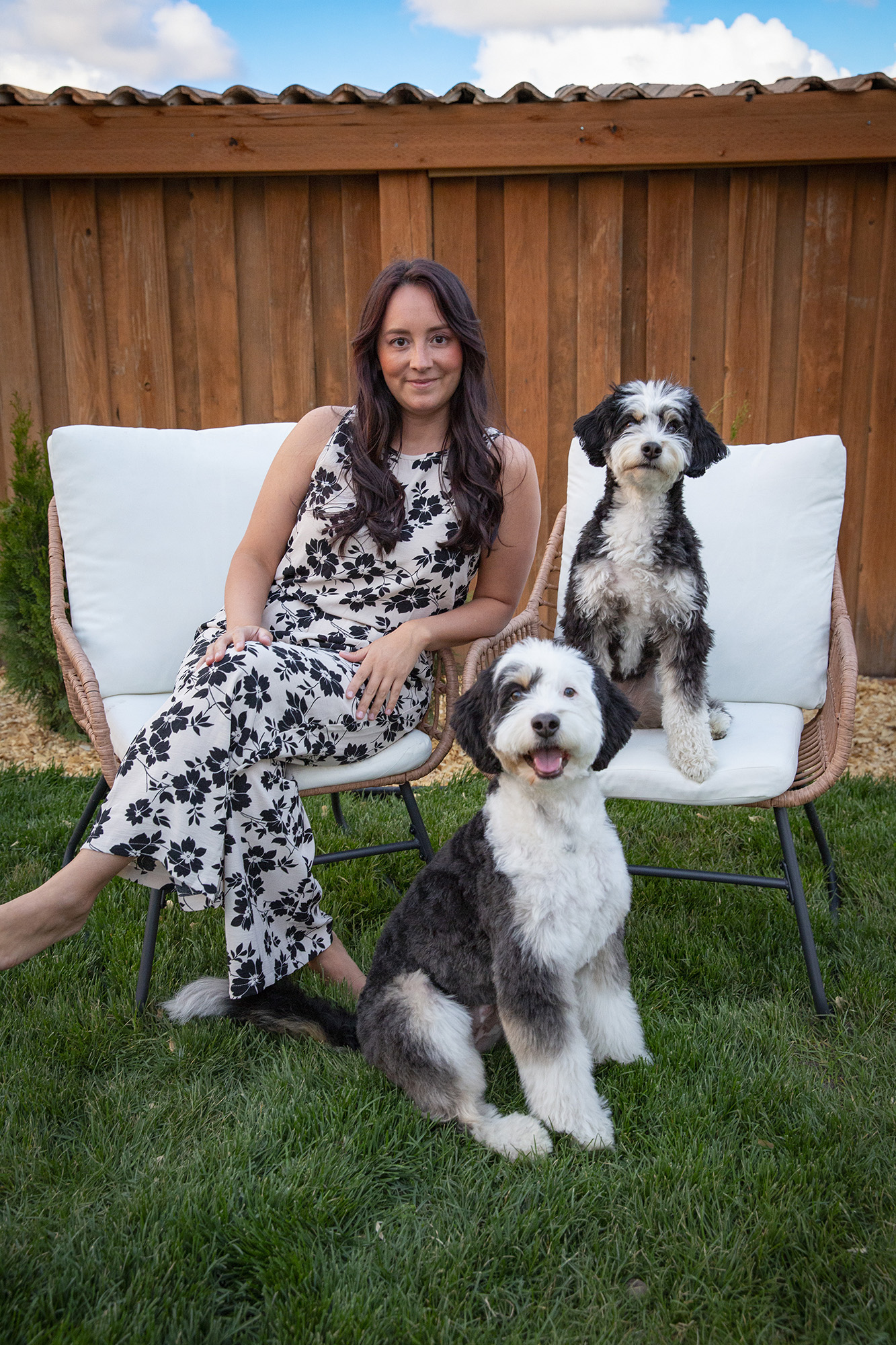 Elaine with her two dogs sitting on wicker chairs with white cushions, with the brown fence behind them and the green grass below. 