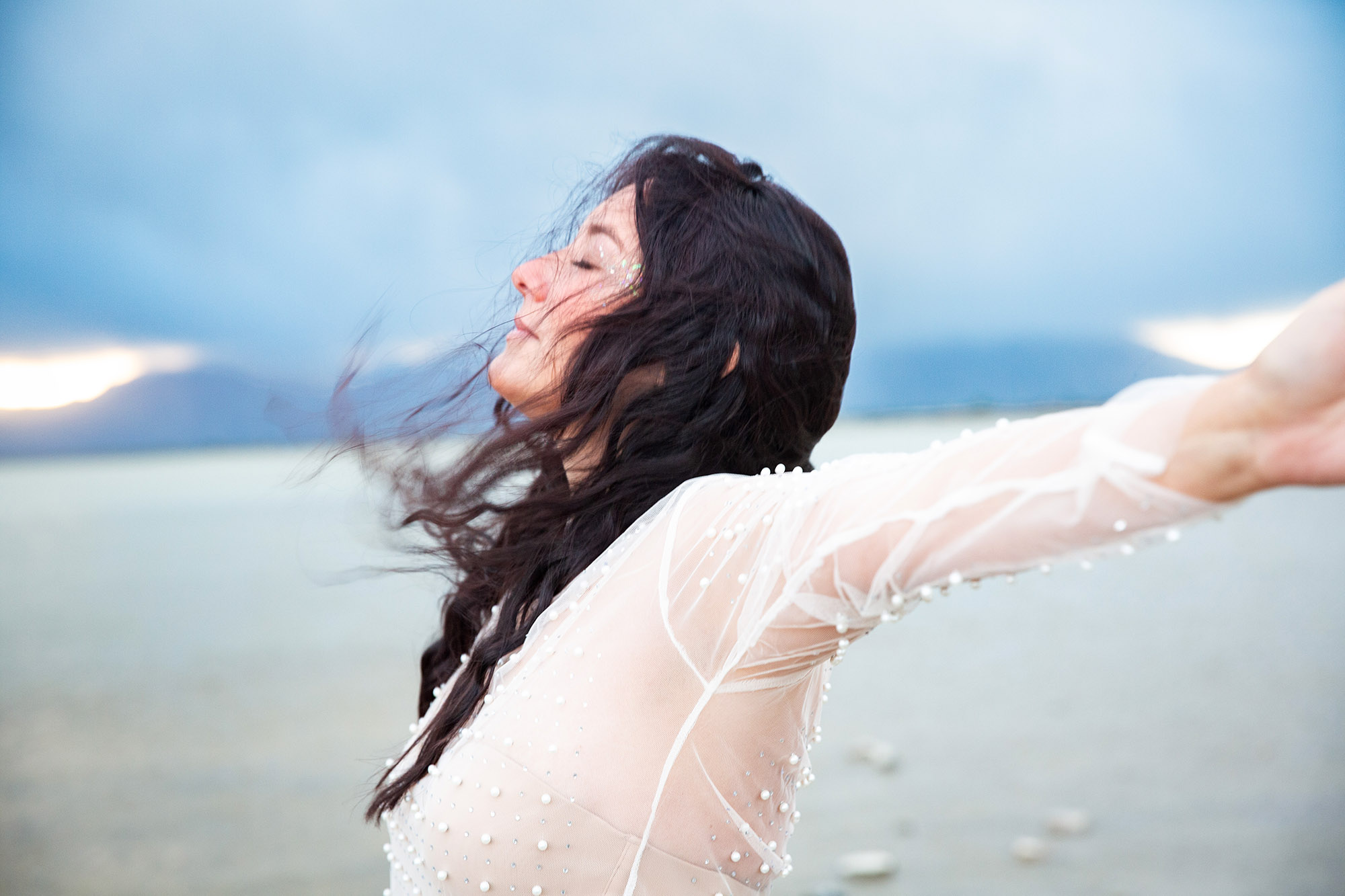 Elaine- woman with long dark hair blowing in the wind with her arms out to the side. Light blue sunrise sky. 