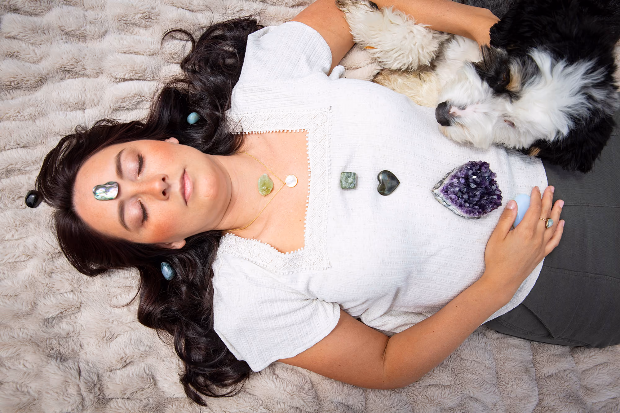 Elaine - woman laying on cream blanket with shells and crystals placed on chakras and black and white dog with it's head on her stomach