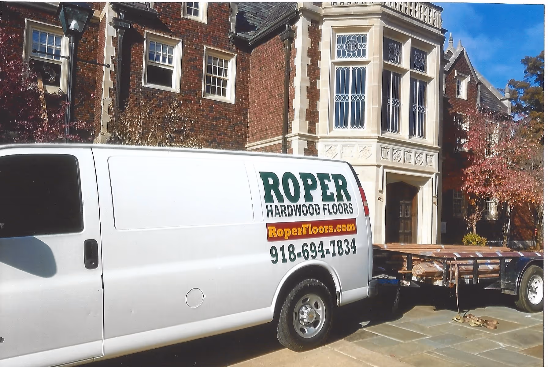 White van labeled 'Roper Hardwood Floors' with phone number and website, parked in front of a large brick building with ornate windows, towing a trailer loaded with wooden planks.