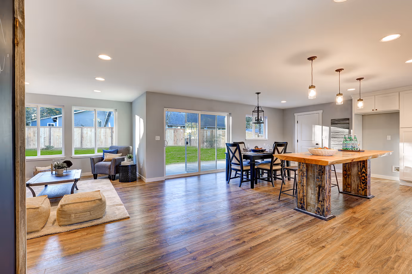Bright open living and dining area with wooden flooring, a rustic wooden dining table, black dining chairs, armchair near windows, and sliding glass doors opening to a yard.