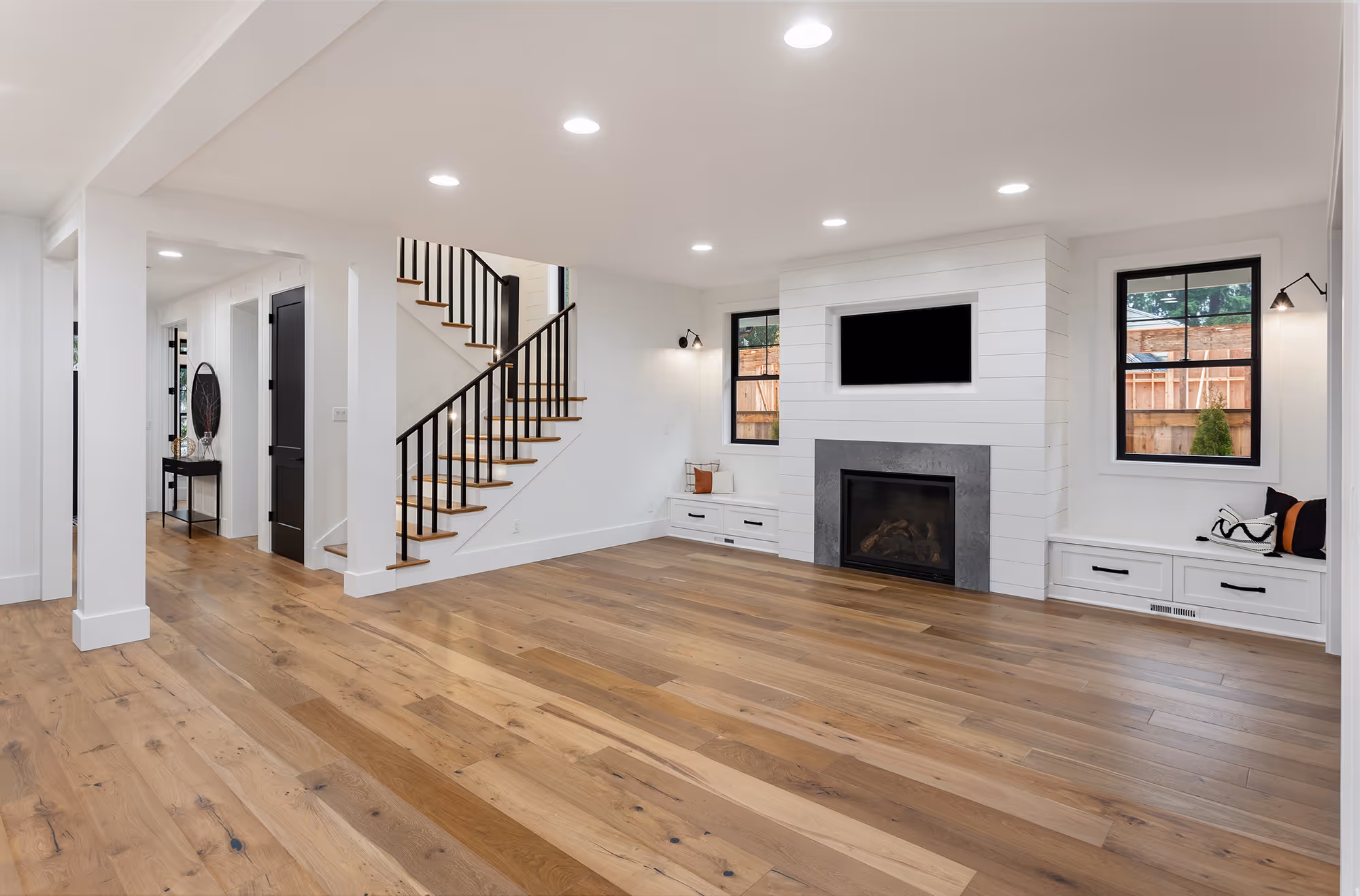 Spacious modern living room with wooden floor, white walls, staircase with black railing, fireplace, and built-in benches under two windows.