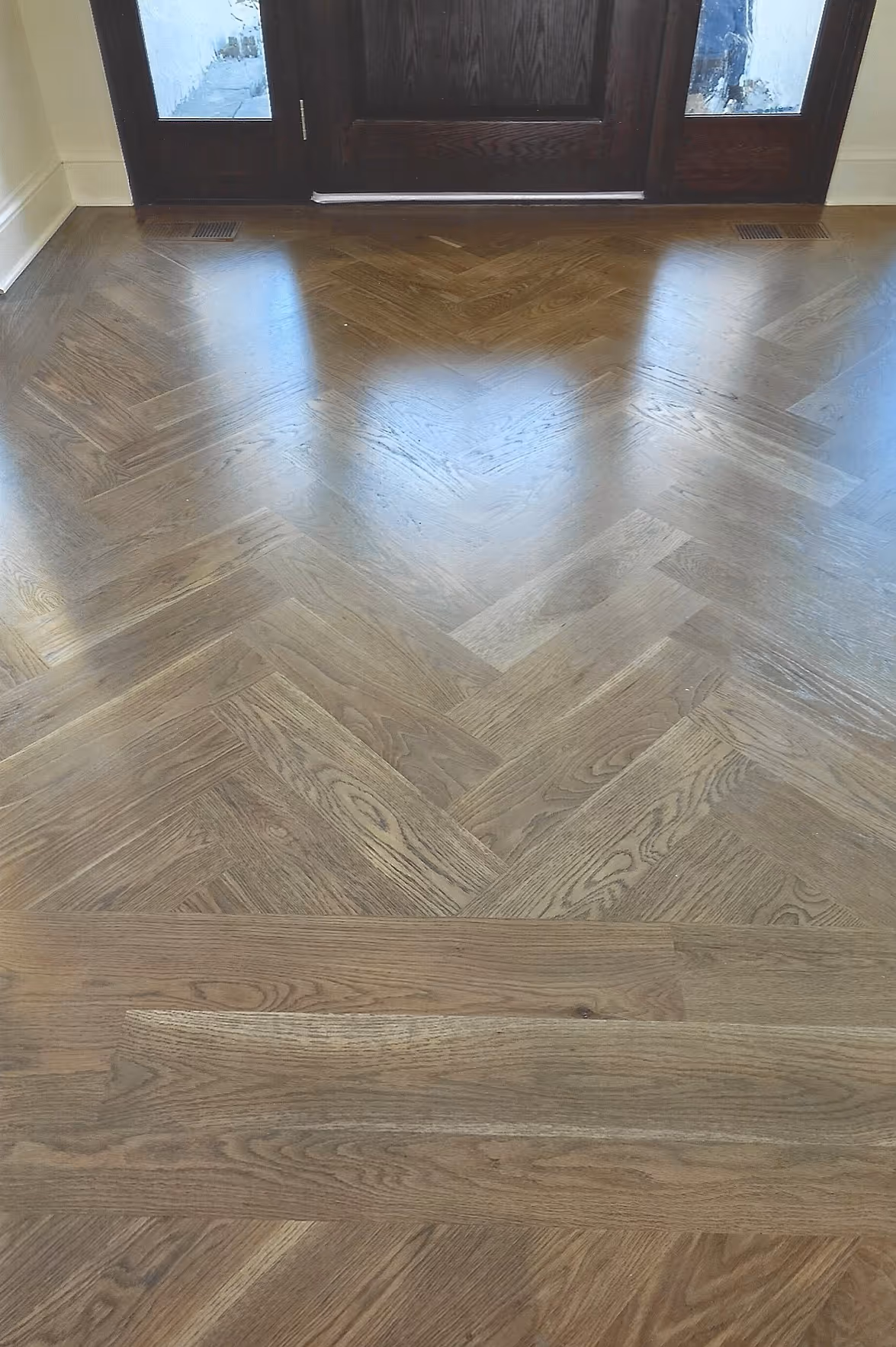 Glossy herringbone-patterned wooden floor with a horizontal wood strip near dark brown front door with glass side panels.