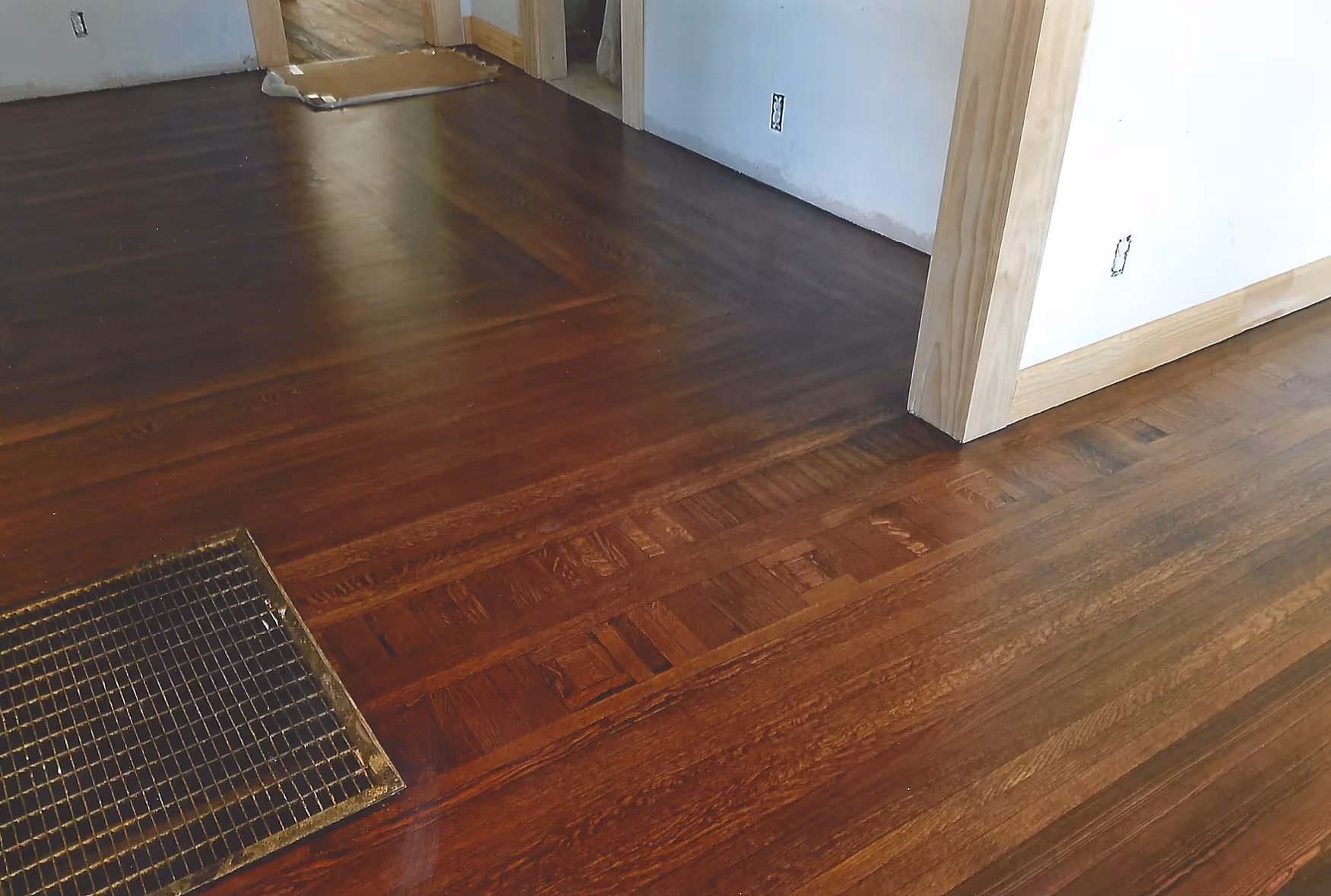 Dark polished hardwood floor with a metal floor vent and unfinished white walls with wooden door frames.