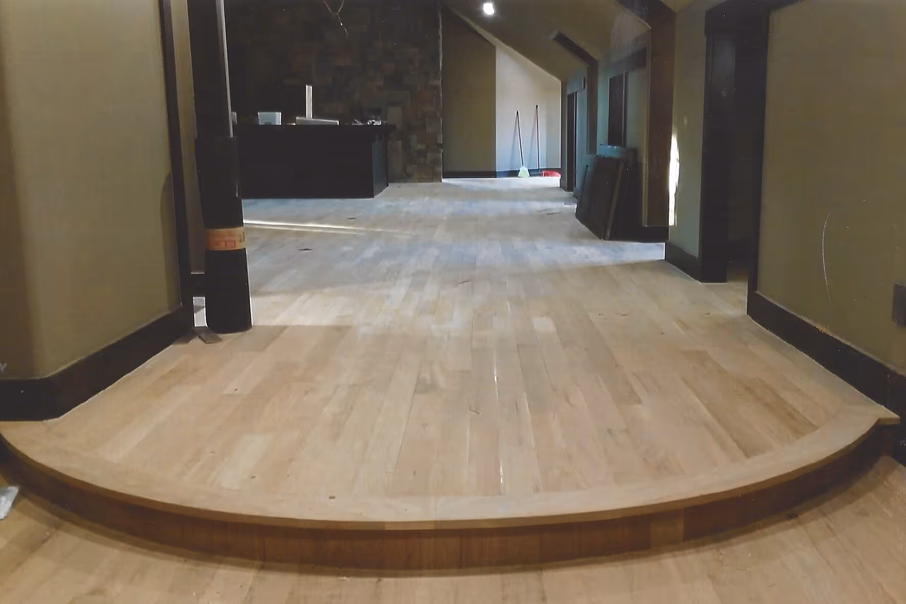 Interior view of an unfinished room with light wood flooring, a curved step, dark trim, and cleaning supplies in the background.