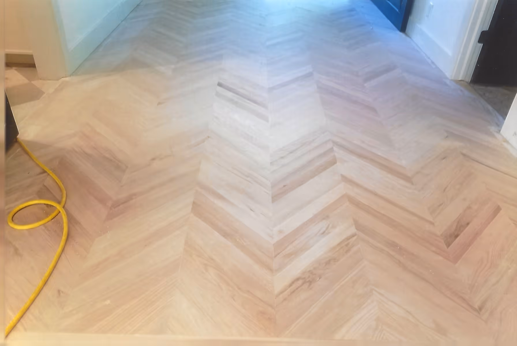 Light wood floor with a herringbone pattern in a hallway, with a yellow extension cord on the left side.