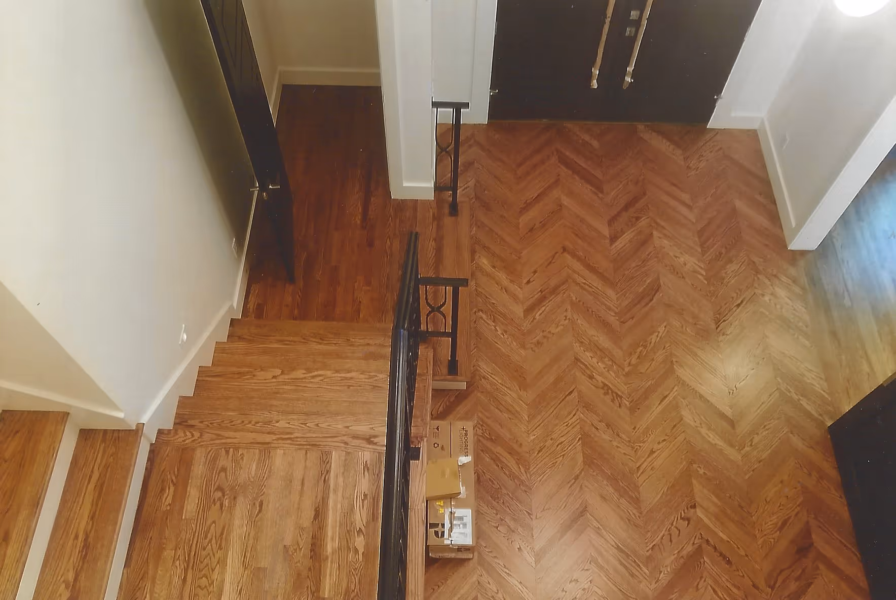 Top-down view of a wooden staircase with black metal railing next to a chevron patterned wooden floor and a closed black door.
