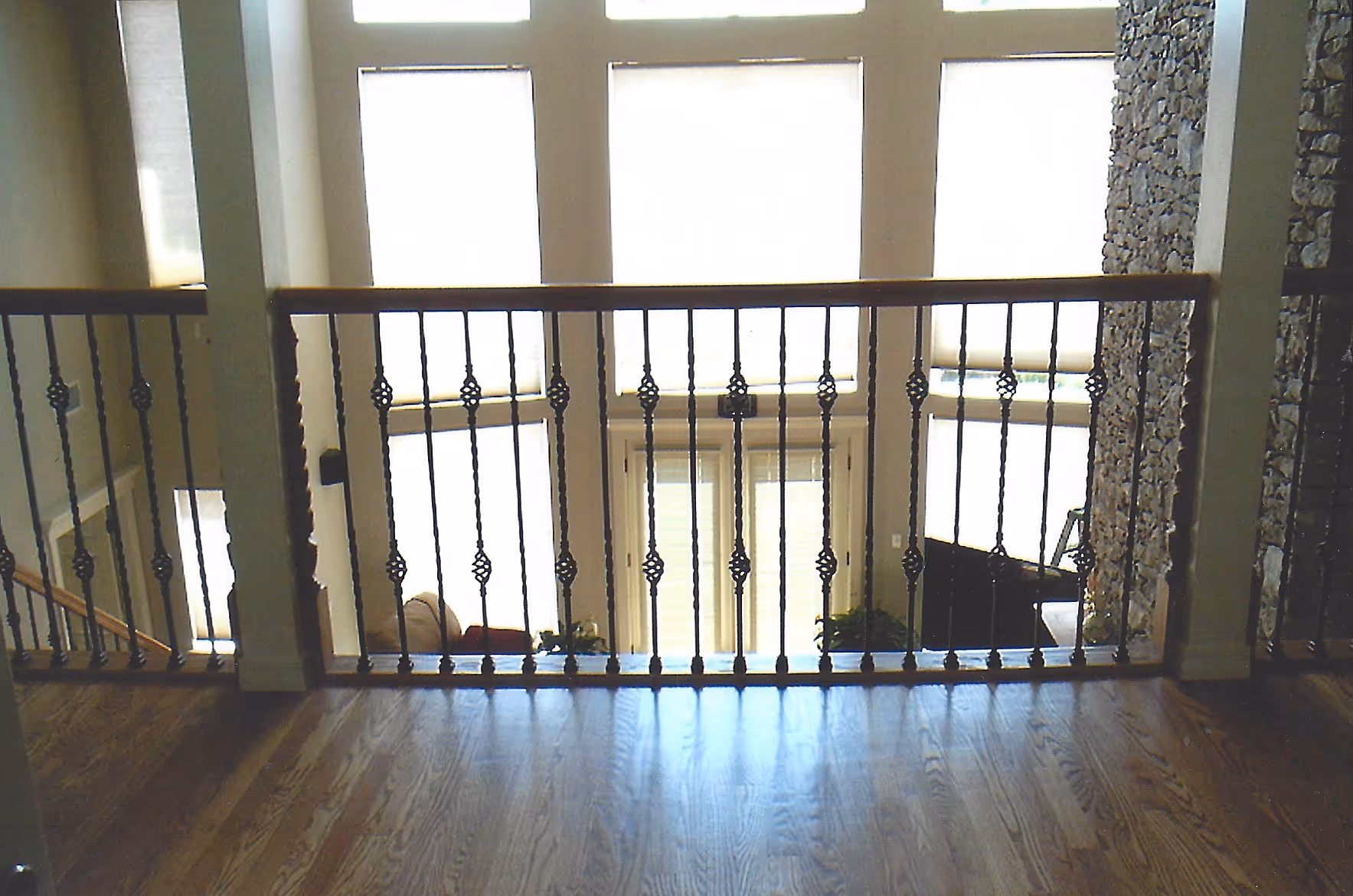 Indoor view from wooden floor balcony with metal railings overlooking a sunlit living room with large windows and stone accent wall.