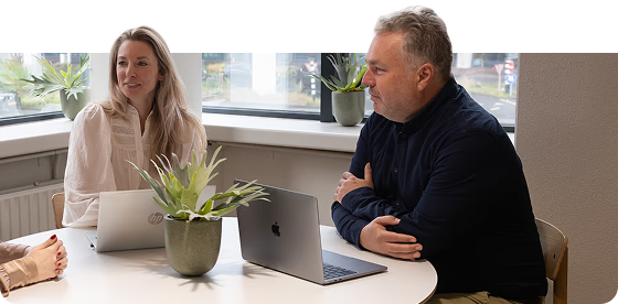 Two colleagues having a business meeting at a round table with laptops and a potted plant near a window.