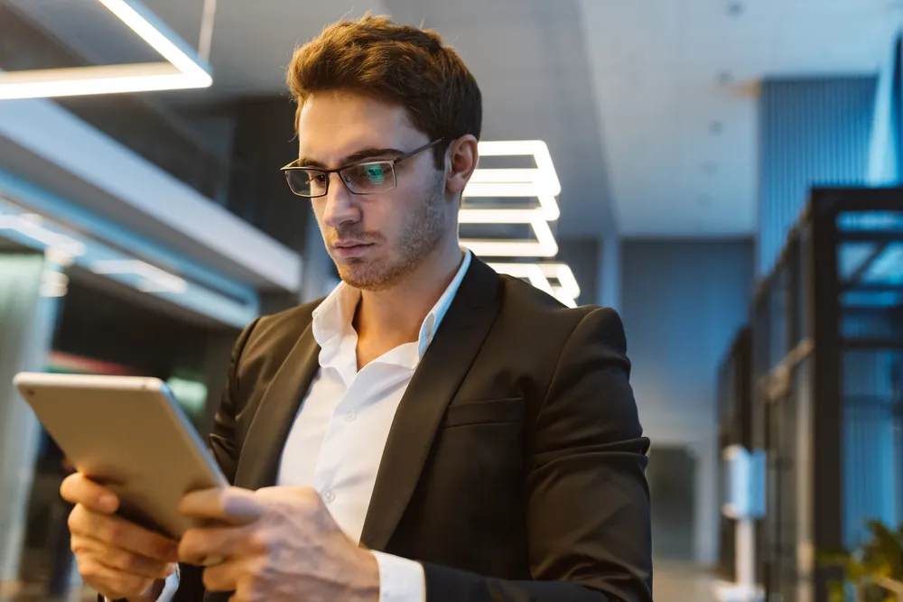 Young man in a black suit and glasses using a tablet in a modern indoor space with geometric ceiling lights.